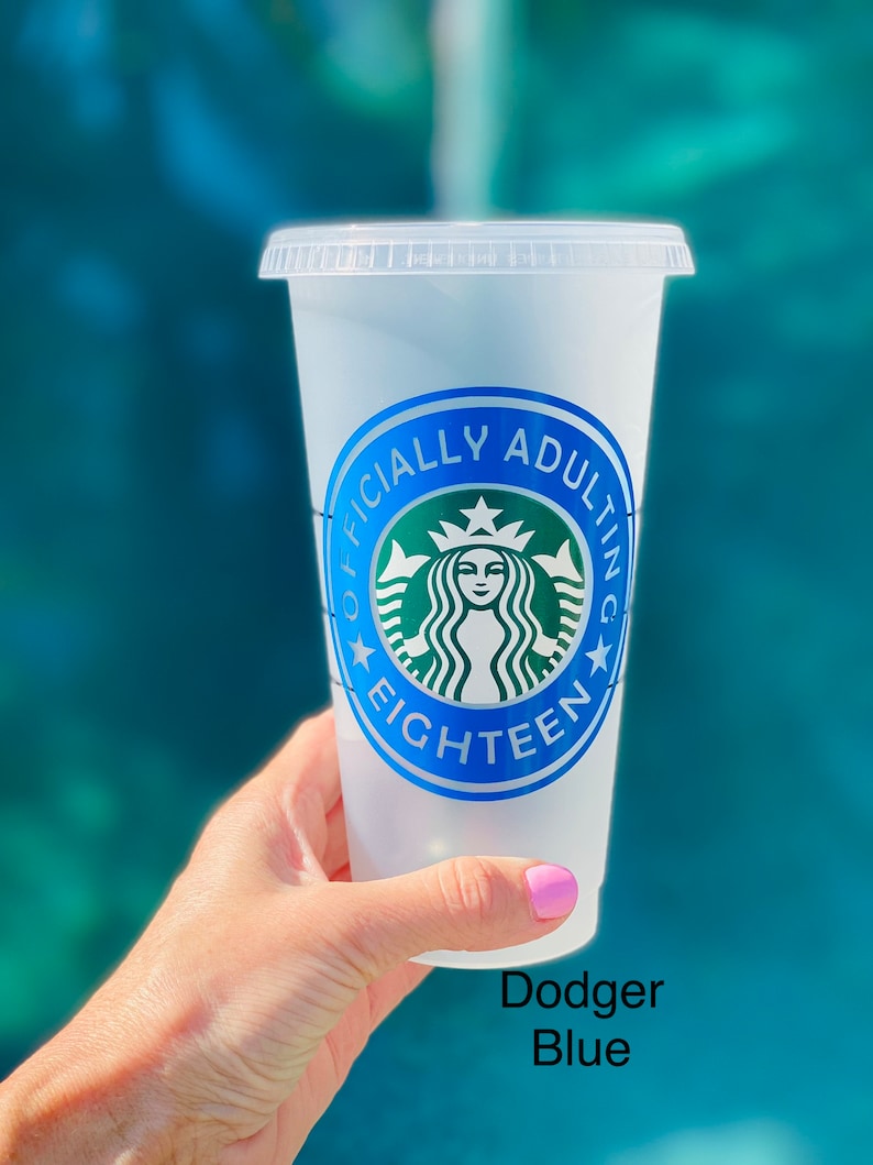 May include: A clear plastic reusable cup with a blue and green sticker that says "Officially Adulting Eighteen" with a stylized green Starbucks logo. The cup is being held by a hand with pink nail polish.