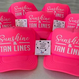Puede incluir: Una colecci&oacute;n de gorras de camionero de color rosa ne&oacute;n brillante con texto serigrafiado en blanco que dice Sunshine & TAN LINES. Cada gorra tiene un panel frontal de espuma, una parte posterior de malla y una visera curva. Peque&ntilde;as tarjetas blancas con el logotipo de Rugged Heart Designs est&aacute;n colocadas entre las gorras.
