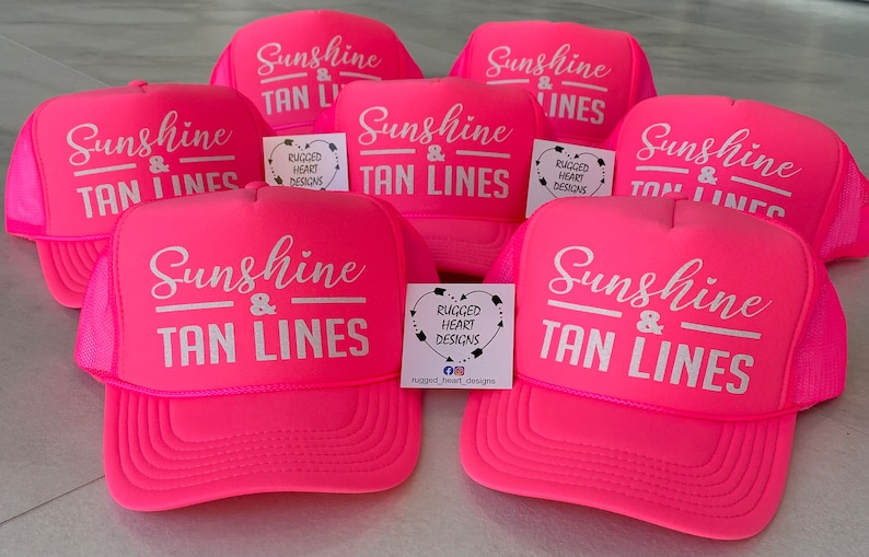 Puede incluir: Una colecci&oacute;n de gorras de camionero de color rosa ne&oacute;n brillante con texto serigrafiado en blanco que dice Sunshine & TAN LINES. Cada gorra tiene un panel frontal de espuma, una parte posterior de malla y una visera curva. Peque&ntilde;as tarjetas blancas con el logotipo de Rugged Heart Designs est&aacute;n colocadas entre las gorras.