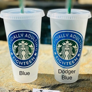 May include: Two blue and white reusable cups with a green Starbucks logo and the text "Officially Adulting Eighteen" on each cup. The cups are sitting on a brown surface with a small white card that says "Rugged Heart Designs".