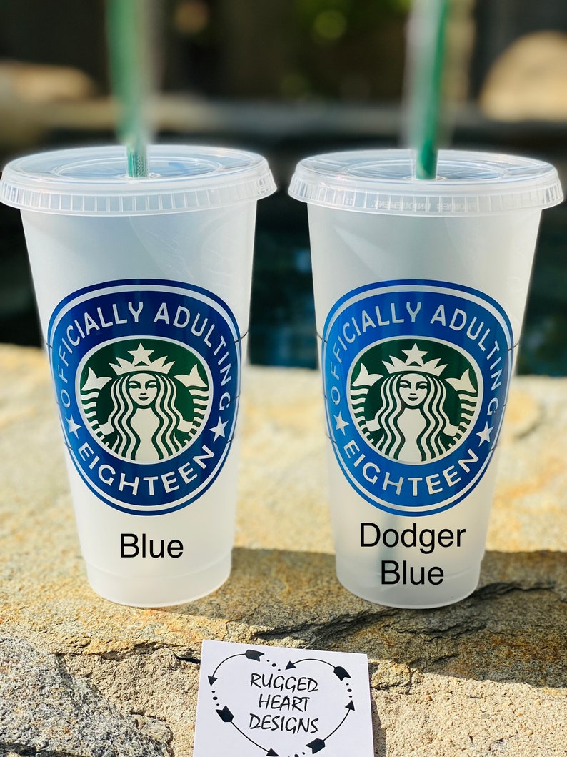 May include: Two blue and white reusable cups with a green Starbucks logo and the text "Officially Adulting Eighteen" on each cup. The cups are sitting on a brown surface with a small white card that says "Rugged Heart Designs".