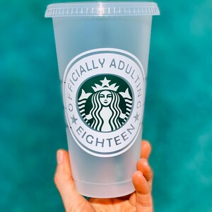 May include: A clear plastic cup with a white sticker that says "Officially Adulting Eighteen" with a green Starbucks logo in the center.