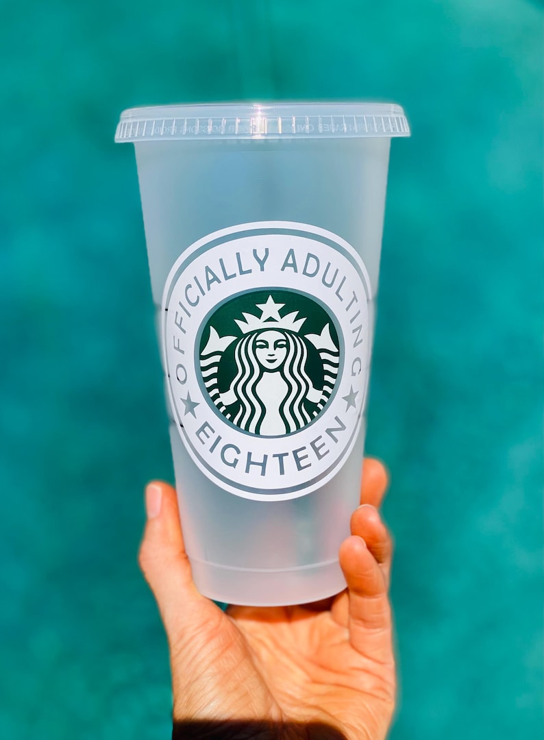 May include: A clear plastic cup with a white sticker that says "Officially Adulting Eighteen" with a green Starbucks logo in the center.