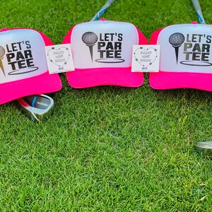 May include: Three pink trucker hats with white fronts, each featuring the text "LET'S PAR TEE" and a golf ball graphic. The hats are on green grass, with golf clubs visible. Each hat has a tag that says "RUGGED HEART DESIGNS".