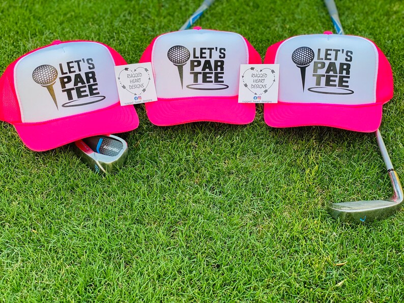 May include: Three pink trucker hats with white fronts, each featuring the text "LET'S PAR TEE" and a golf ball graphic. The hats are on green grass, with golf clubs visible. Each hat has a tag that says "RUGGED HEART DESIGNS".