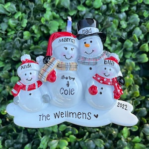 May include: A festive Christmas ornament depicting a snowman family. The ornament features snowmen with the names "Andi", "Ella", "Mama", "Dad", and "Baby Cole". The ornament is white with red and black details, and the year "2022" is displayed.