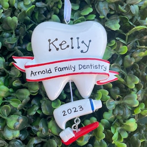 May include: White tooth-shaped Christmas ornament with a red ribbon that says "Arnold Family Dentistry". The tooth has the name "Kelly" written on it and a red and white toothbrush with a tube of toothpaste that says "2023" hanging from it.