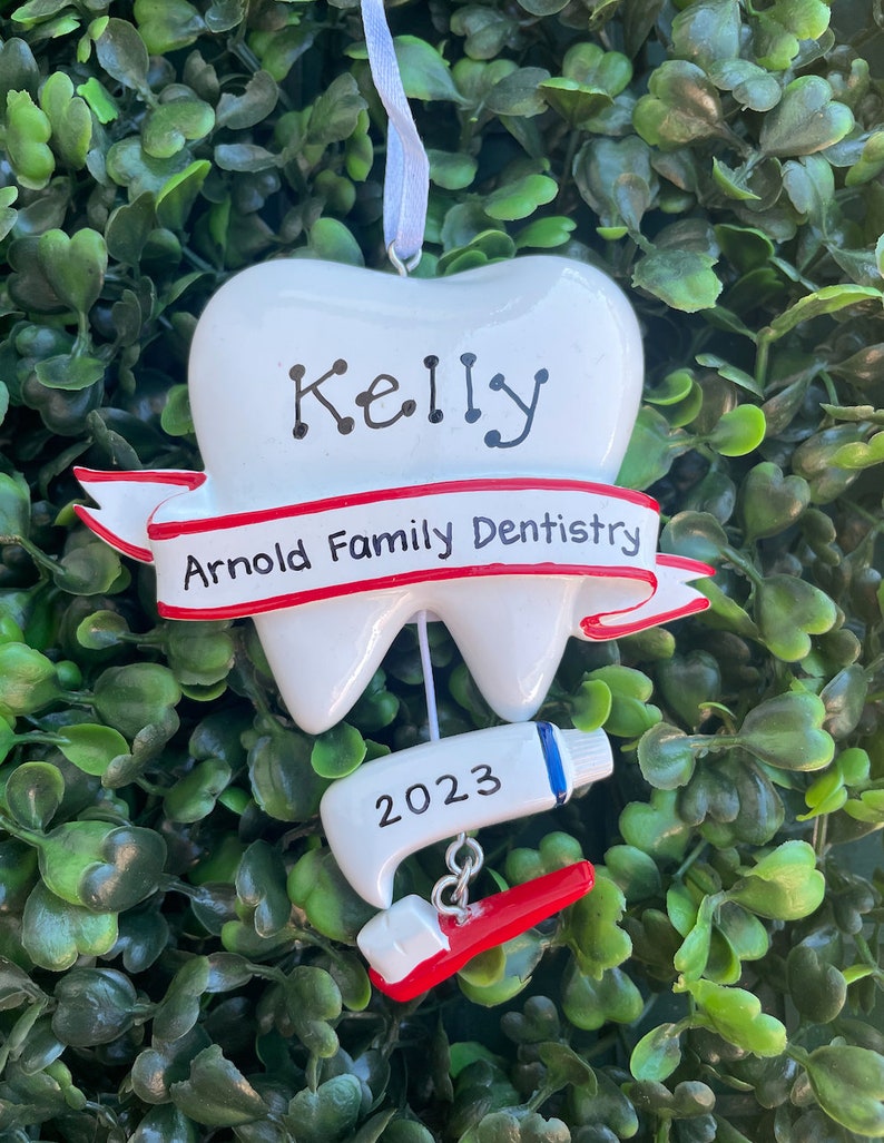 May include: White tooth-shaped Christmas ornament with a red ribbon that says "Arnold Family Dentistry". The tooth has the name "Kelly" written on it and a red and white toothbrush with a tube of toothpaste that says "2023" hanging from it.