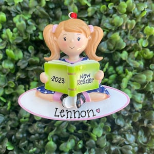 May include: A personalised ornament depicting a cartoon girl with blonde pigtails reading a book. The book reads "New Reader 2023". The ornament is on a white oval base with the name "Lennon" and a heart.