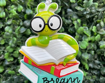 Op de afbeelding: Een groen en geel rupsornament met een bril, bovenop een stapel boeken. De boeken zijn rood, geel en turquoise, met de naam "Breann" en het jaar "2025" zichtbaar. Het ornament heeft een zilveren lus om op te hangen.