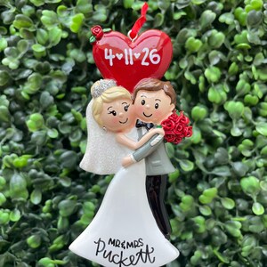 May include: A wedding ornament featuring a bride and groom figurine embracing. The bride wears a white dress and veil, while the groom wears a gray suit. Above them is a red heart with the date 4-11-26. The base reads Mr & Mrs Puckett.