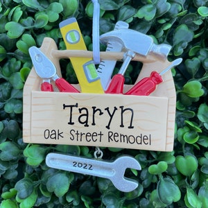 May include: A wooden toolbox ornament with various tools, including a level, hammer, and screwdriver. The ornament has the name "Taryn" and "Oak Street Remodel" printed on it. A silver wrench with "2022" is attached.