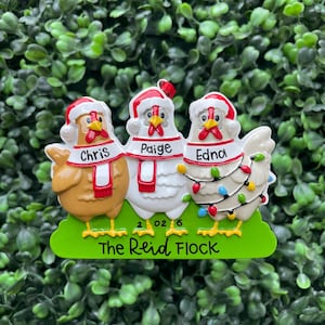 May include: A Christmas ornament featuring three chickens wearing Santa hats and red and white scarves. The chickens are named Chris, Paige, and Edna. The ornament includes a string of colorful lights, the year 2026, and the text The Reid Flock.