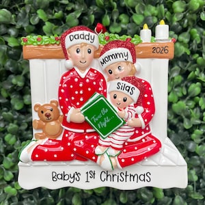 May include: A Christmas ornament featuring a family of three in red and white polka dot pajamas sitting by a fireplace. The figures wear hats labeled Daddy, Mommy, and Silas. A green book titled Twas the Night is held, with a teddy bear and 2026 text.