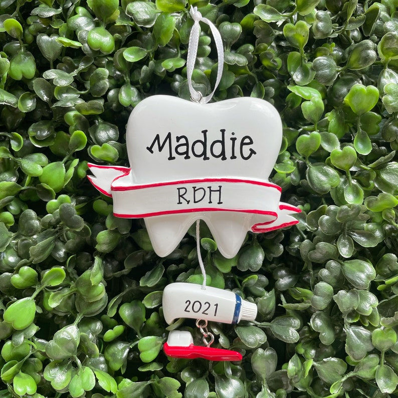 May include: White tooth-shaped Christmas ornament with a red ribbon that says "Maddie RDH" and a white tube of toothpaste with "2021" written on it.