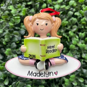 May include: A hanging ornament featuring a light-skinned child with blonde pigtails reading a lime green book labeled New Reader and 2026. The child wears a blue dress with white polka dots and sits on a white oval base inscribed with Madelyn.