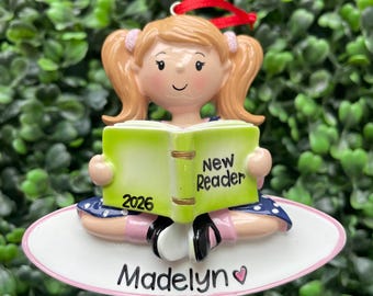 Op de afbeelding: Een hangend ornament met een lichtgetint kind met blonde staartjes dat een limoengroen boek leest met het opschrift New Reader en 2026. Het kind draagt een blauwe jurk met witte stippen en zit op een witte ovale basis met de naam Madelyn.