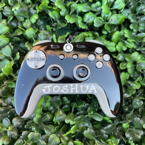 May include: Black and grey video game controller ornament with the year 2023 and the name Joshua printed on it.