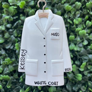 May include: A white lab coat ornament featuring a collar, five black buttons, and two front pockets. The text MUSC is printed on the chest pocket, Kelsey is on the right sleeve, 2026 is on the bottom right, and WHITE COAT is at the bottom hem.
