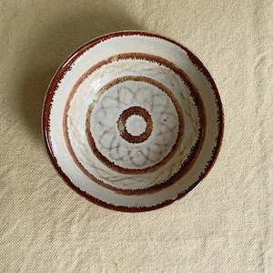 May include: A white ceramic bowl with brown concentric circles and a brown floral design in the centre.