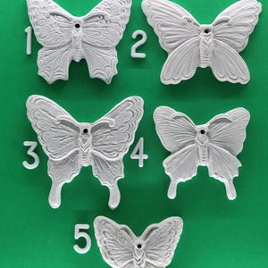 May include: Five white butterfly-shaped decorative items are arranged on a green surface. Each butterfly has a unique wing pattern and is numbered from one to five. The butterflies are made of a solid material and have a detailed, textured appearance.