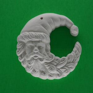 May include: A white, unpainted ceramic Santa Claus head ornament in a crescent moon shape. The ornament features a detailed face, beard, and hat, with a hole at the top for hanging. The background is a solid green.
