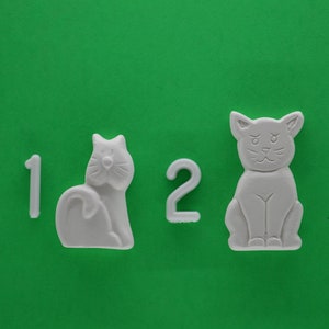 May include: Two white ceramic cat figurines with the numbers 1 and 2 on a green background.