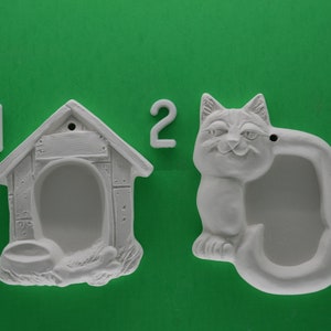 May include: Two white ceramic ornaments, one shaped like a dog house with a food bowl and the other shaped like a cat with a curled tail. The ornaments are numbered 1 and 2.