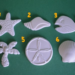 May include: Six white ceramic ornaments with a beach theme. The ornaments are numbered 1 through 6. The ornaments include a starfish, a seashell, a whale, a palm tree, a sand dollar, and a scallop shell.