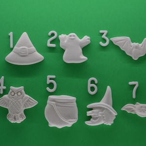 May include: Seven white ceramic Halloween-themed ornaments, numbered 1 through 7. The ornaments include a witch's hat, a ghost, a bat, an owl, a cauldron, a witch, and another bat.