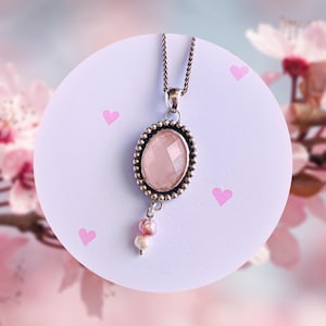 Könnte beinhalten: Eine silberne Halskette mit einem ovalen, facettierten Anhänger aus rosa Edelstein. Der Anhänger ist von einem Perlenrand umgeben und hat eine kleine Ansammlung von rosa und weißen Perlen, die darunter hängen. Die Halskette wird auf einem weißen Hintergrund mit rosa Herzakzenten präsentiert.