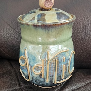 May include: A green and blue ceramic jar with a lid. The jar has the word "garlic" written on it in a light brown glaze.