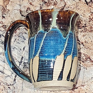 May include: A handmade ceramic mug with a blue, brown and white glaze. The mug has a unique, abstract design with drips and streaks of colour.