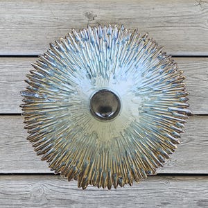 May include: A circular ceramic wall plate featuring a sunburst pattern with radiating spikes. The center is a smooth, dark brown circular indentation, while the surrounding surface transitions from a light cream to a metallic bronze and blue glaze.