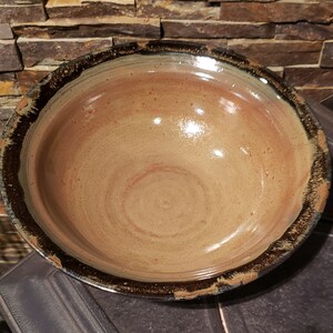May include: A handcrafted ceramic bowl with a wide, shallow form. The interior displays warm, earthy hues of tan and reddish-brown, with a dark, textured brown rim. The bowl's surface has a glossy finish, reflecting light.