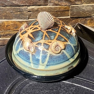 May include: A blue and brown ceramic butter dish with a seashell design. The lid features a seashell, a snail, and two crab claws.