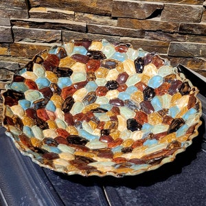 May include: A large ceramic bowl with a scalloped edge. The bowl is decorated with a mosaic of small, irregularly shaped tiles in shades of brown, blue, and cream.