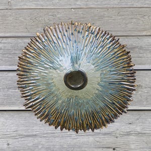 May include: A circular ceramic wall plate featuring a sunburst pattern with radiating spikes. The glaze transitions from a dark brown centre to a blend of light blue, green, and tan tones along the textured, pointed edges of the piece.