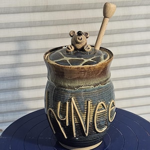 May include: A ceramic honey pot with a bear figurine on the lid and the word "Honey" written on the side. The pot has a brown, blue, and white glaze and comes with a wooden honey dipper.