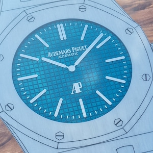 Könnte beinhalten: Ein silberfarbenes Zifferblatt mit einem blauen Zifferblatt und weißen Markierungen. Das Zifferblatt der Uhr zeigt "Audemars Piguet Automatic" und "AP".