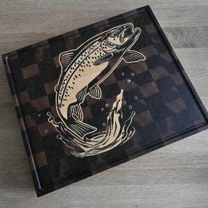 Könnte beinhalten: Quadratisches Holzschneidebrett mit einem dunkelbraunen und beigefarbenen Schachbrettmuster. Eine detaillierte Illustration eines Fisches, der aus dem Wasser springt, ist in das hellfarbige Holz geschnitzt. Das Brett hat eine vertiefte Kante.