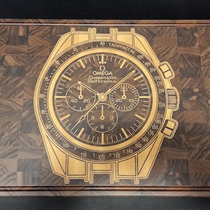 Könnte beinhalten: Ein Schneidebrett aus Holz mit einem goldfarbenen Bild einer Omega Speedmaster Professional Chronographenuhr, das in die Oberfläche graviert ist.