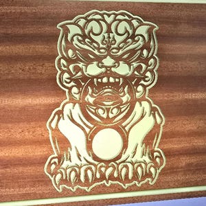 Könnte beinhalten: Holztafel mit einem detaillierten, geschnitzten Design einer mythischen Kreatur. Die Kreatur ist goldfarben, vor einem braunen Holzmaserungshintergrund. Die Tafel hat einen hellfarbigen Rand.