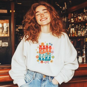Könnte beinhalten: Weißes Sweatshirt mit dem Wort "MAMA" in einem Retro-Schriftzug, umgeben von bunten Blumen und Smiley-Gesichtern. Das Sweatshirt wird mit einer blauen Jeans kombiniert. Der Gesamtstil ist eine Vintage- und Groovy-Ästhetik.