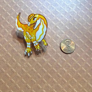 May include: A yellow and white cartoon dinosaur pin with a toothy grin. The pin is about the size of a US penny.