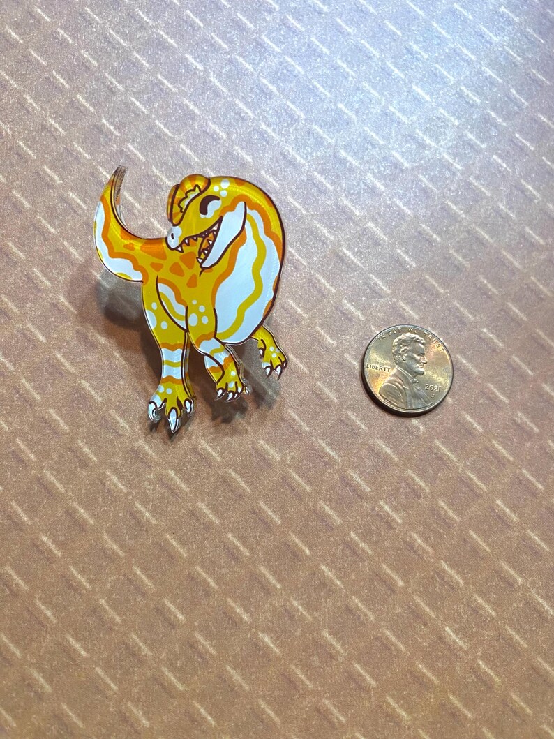 May include: A yellow and white cartoon dinosaur pin with a toothy grin. The pin is about the size of a US penny.