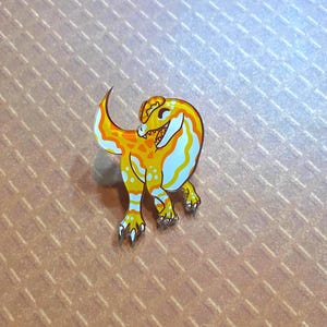 May include: A yellow and white enamel pin of a cartoon dinosaur with a toothy grin. The dinosaur is standing on its hind legs with its tail curled up behind it.