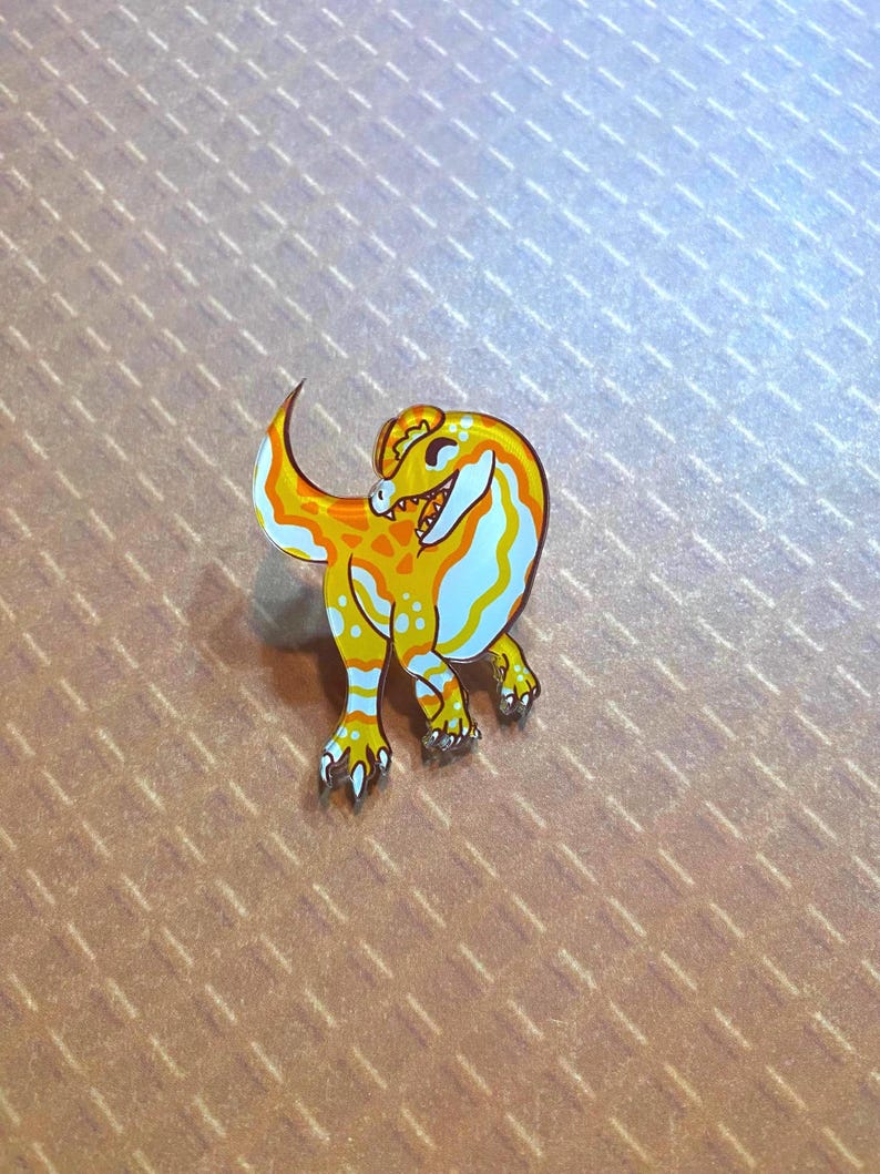 May include: A yellow and white enamel pin of a cartoon dinosaur with a toothy grin. The dinosaur is standing on its hind legs with its tail curled up behind it.