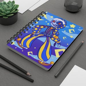 May include: Spiral-bound notebook featuring a cartoon character with a crescent moon face, star-patterned clothing, and yellow and blue striped pants. The background is blue with yellow stars and geometric shapes. Two pencils and a small succulent plant are also visible.