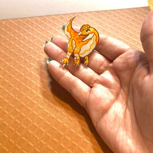 May include: A yellow and orange enamel pin of a cartoon dinosaur with a toothy grin. The dinosaur is standing on its hind legs with its tail curled up.